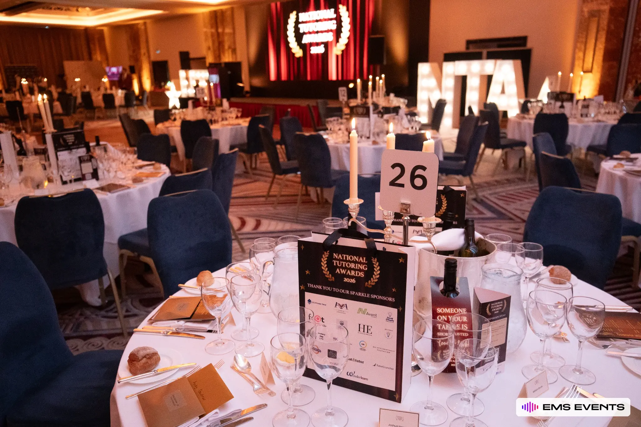 Close-up of a guest table at the 5th National Tutoring Awards with programme, sponsor bag and stage visible behind