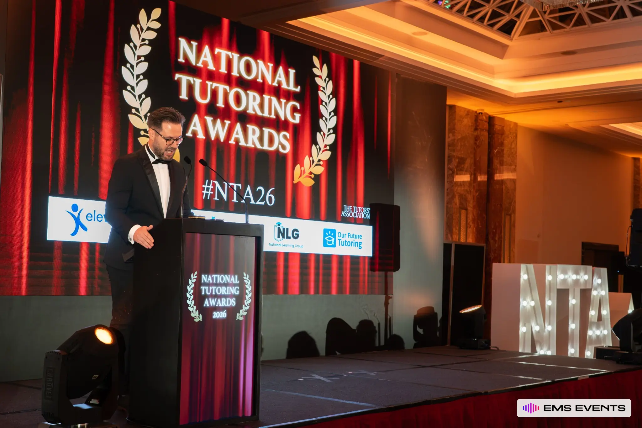 Host addresses guests from the podium with the National Tutoring Awards backdrop and #NTA26 hashtag on the LED screen